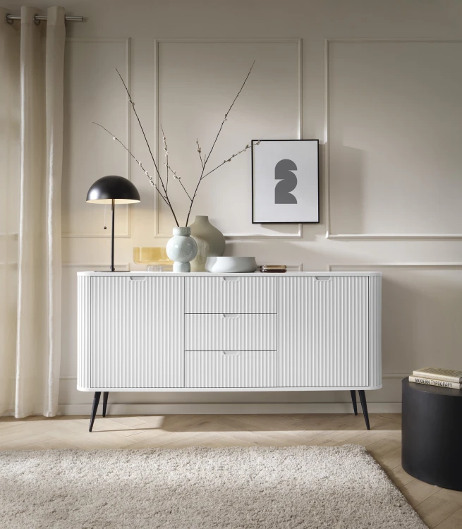 Biała komoda szeroka Zova KSZ168 w aranżacji jasnego salonu, minimalistyczny design z czarnymi akcentami na wysokich nogach